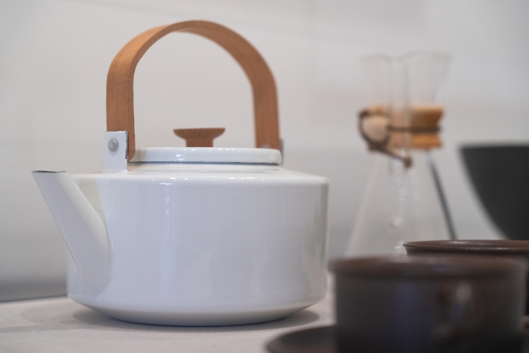 A white enameled tea kettle with a rounded wooden handle. Out of focus and partially showing are a Chemex coffee maker and two dark brown mugs.