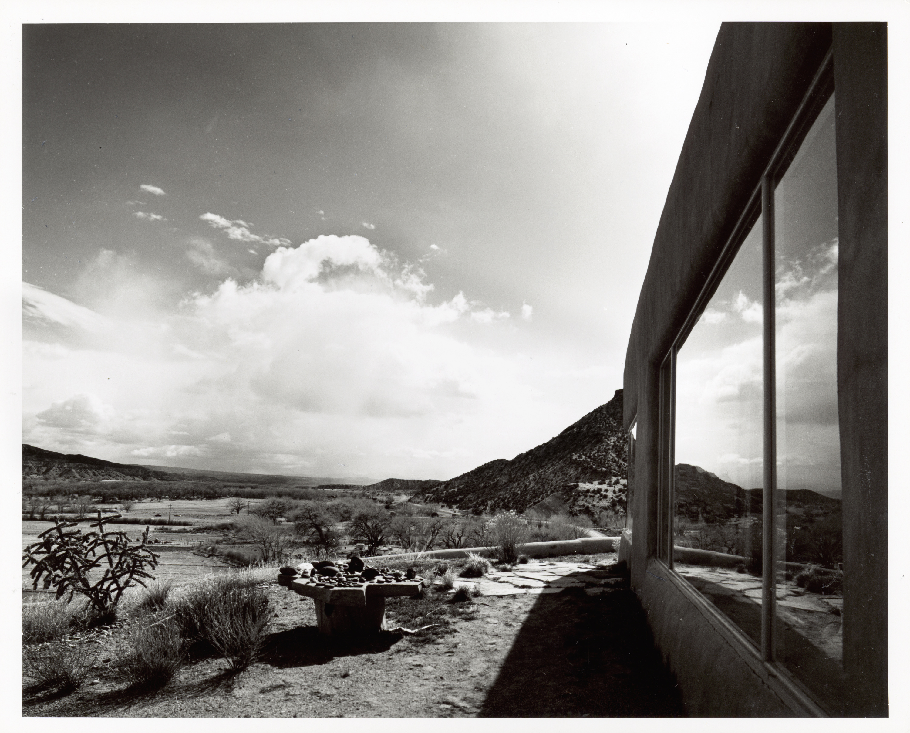 A black and white photo showing a valley from above against the suggestion of a moutnain. An exterior wall in the foreground with a large window reflects rounded mountains in the distance.