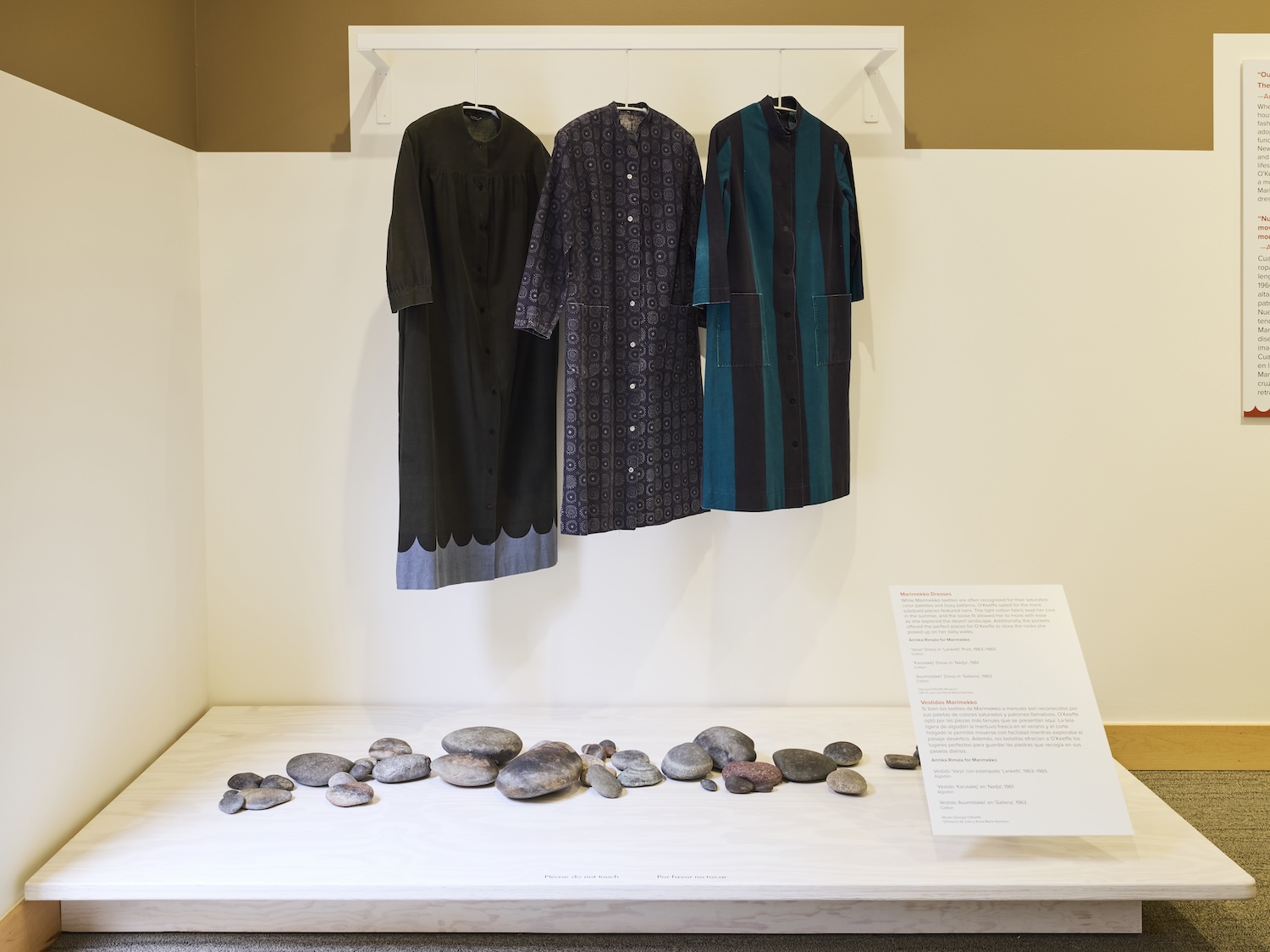 Three dresses hang but face forward against a white wall with a tan outline. The first dress is black with a grey scallopped pattern at the hem. The second has a subtle pattern in gray and black and the third has wide verticle stripes of black and teal. Under the dresses are a pile of smooth round rocks and an unreadable label.