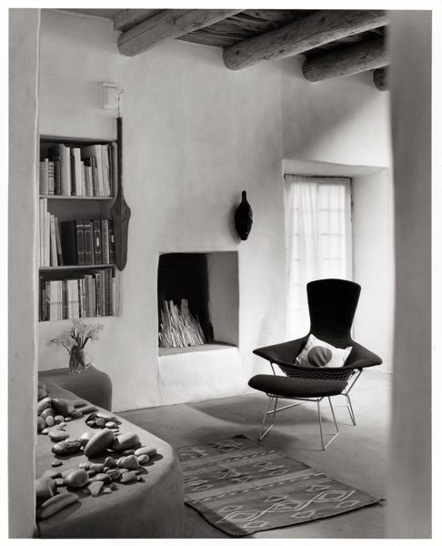 A black and white photography of a living room setting features a dark upholstered chair with organice curves supported by a metal structure. There is a pillow with a circle sitting in the seat and a matching ottoman. The chair sits next to a built in fireplace a window and built-in shelves. The foreground shows the edge of a ledge covered in rounded rocks.