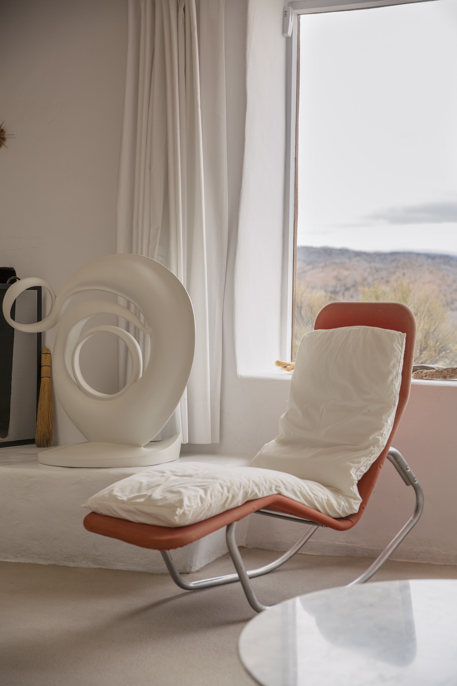 An orange lounger chair with a white cushion faces the interior of a room next to a white abstract circular sculpture in front of a window.