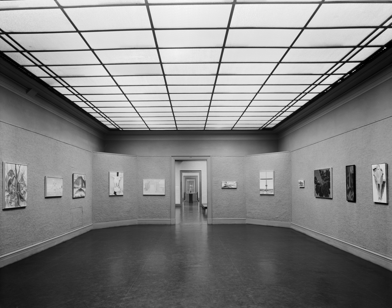 Black and white photograph. This room has an opening at the far end, and walls angle inward across the corners to either side of it. Ten paintings are hung along a textured wall that appears gray in this photograph. The paintings show abstracted or stylized trees, mountains, skulls, or layered shapes. With the grid of lights above and the dark floor below, we look into at least three more rooms, barely visible to either side of the nested doorways.