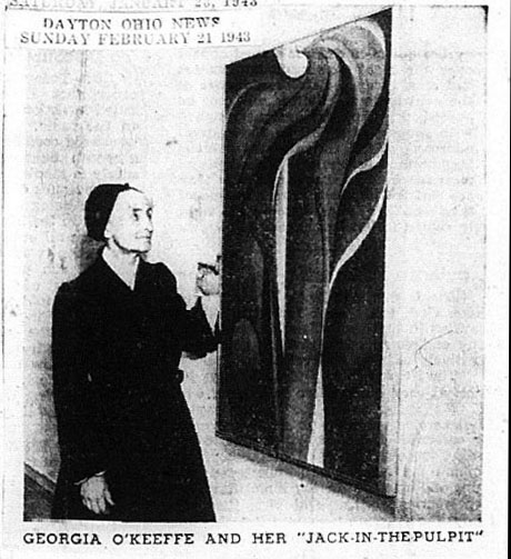 Black and white newspaper clipping. In this grainy image, O’Keeffe stands looking up at a painting of flame-like, flaring petals. The caption beneath reads, 'Georgia O'Keeffe and her 'Jack-in-the-Pulpit.'' An inset box at the top left reads, 'Dayton Ohio News Sunday February 21 1943.' O’Keeffe wears a black, long-sleeved dress and a black cap that covers her hair. Her features are indistinct in this blurry image but dark brows stand out on her high forehead. She has a long nose, and she smiles slightly. She rests the pinky edge of one hand along the side of the canvas.