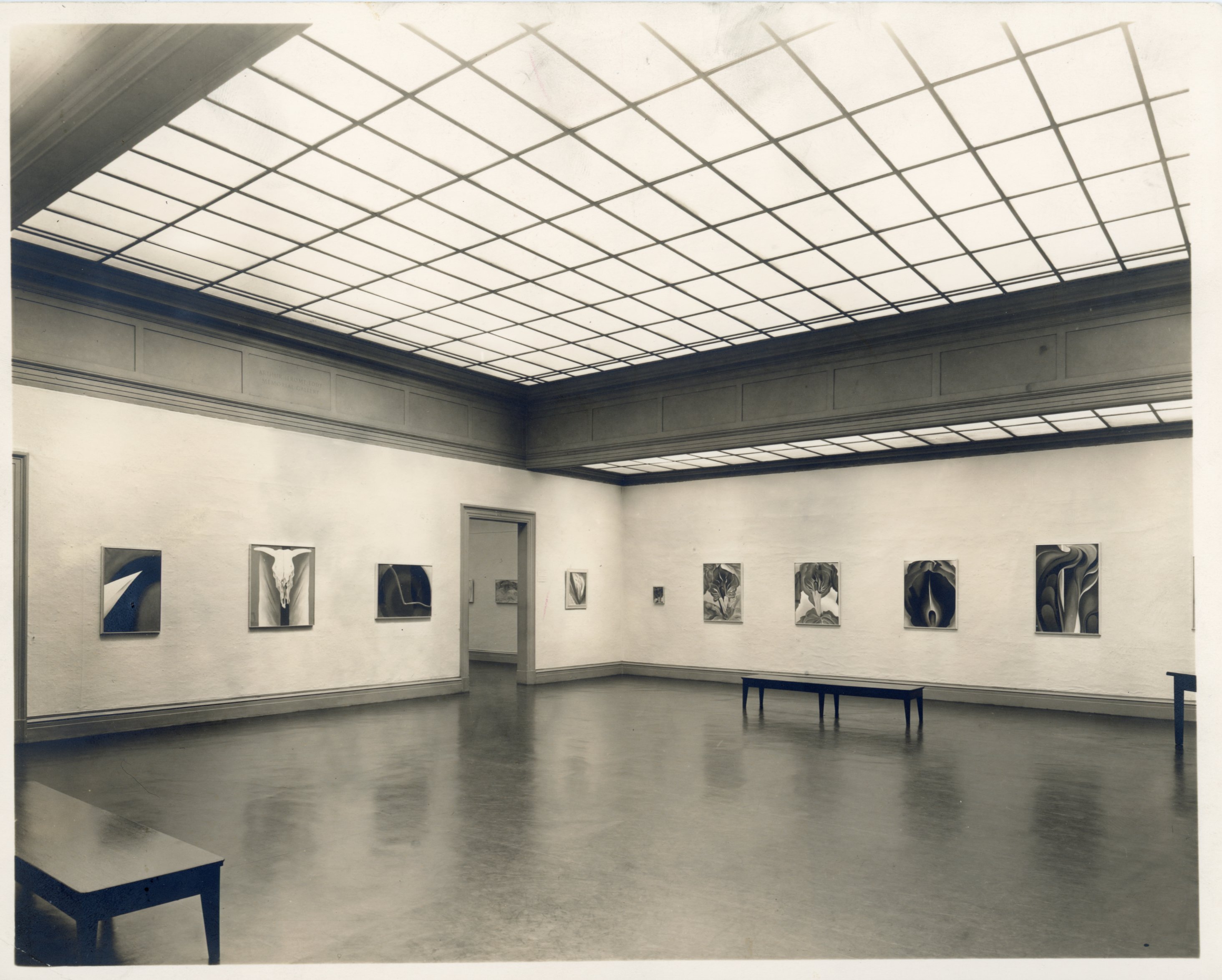 Black and white photograph. We look into the far corner of a gallery space, with nine paintings lining the walls to either side of an opening, near the far corner. The paintings show flame-like petals, an animal skull, or abstracted, geometric shapes. The grid of lights above reflects in the dark, shiny floor below. In this view, we see three wooden benches placed along the perimeter of the room.
