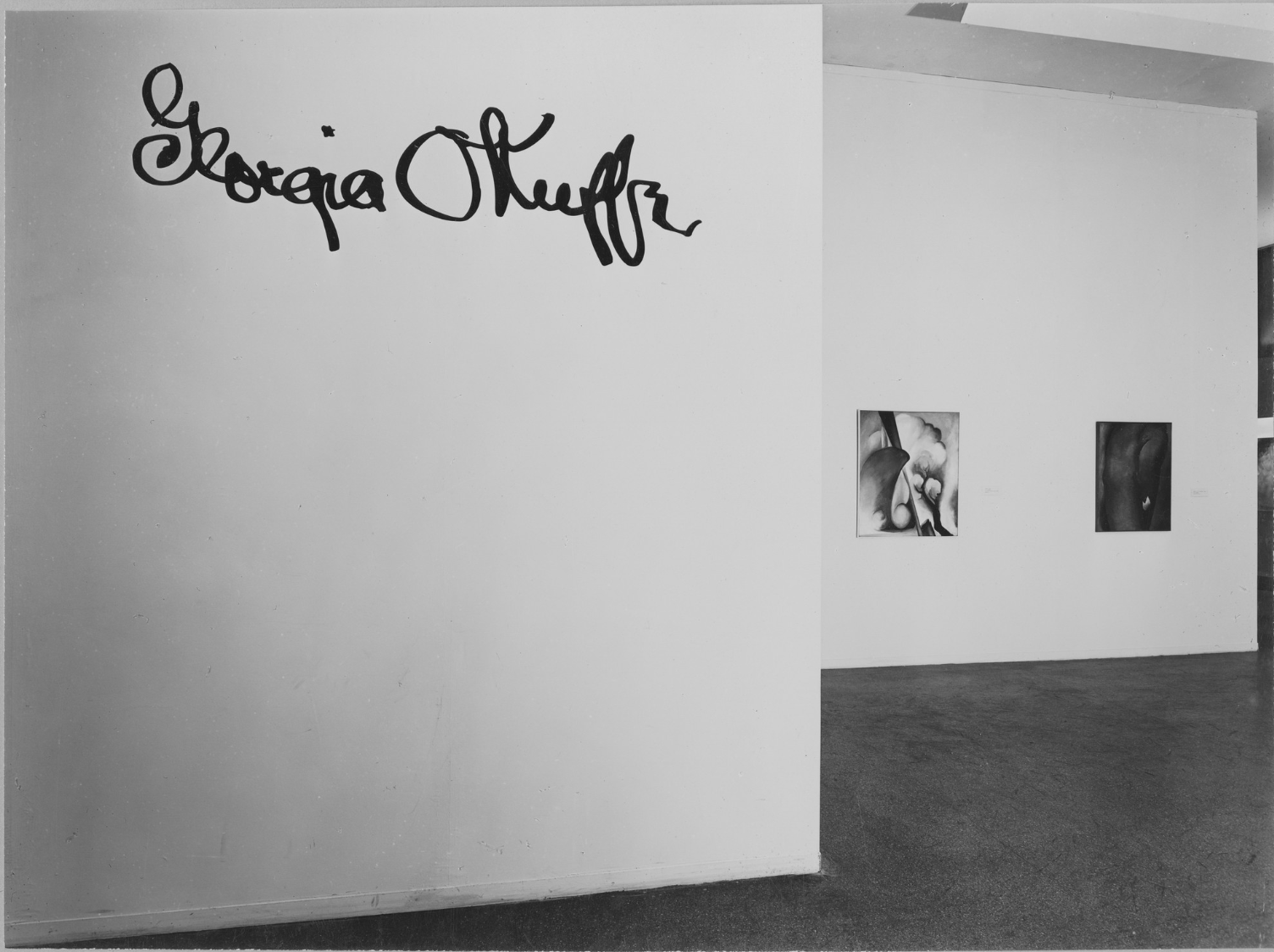 Black and white photograph. A white wall with Georgia O’Keeffe’s signature scrawled in black, oversized, cursive letters, takes up the left half of this image. The room beyond is hung with two paintings, and the sliver of an opening leading to another space is barely visible along the right edge. The letters of O’Keeffe’s name are rounded, with the lowercase g and f letters making spikes along its length. One painting in the background is lighter and shows stylized, curving forms; the other painting is dark and difficult to make out.