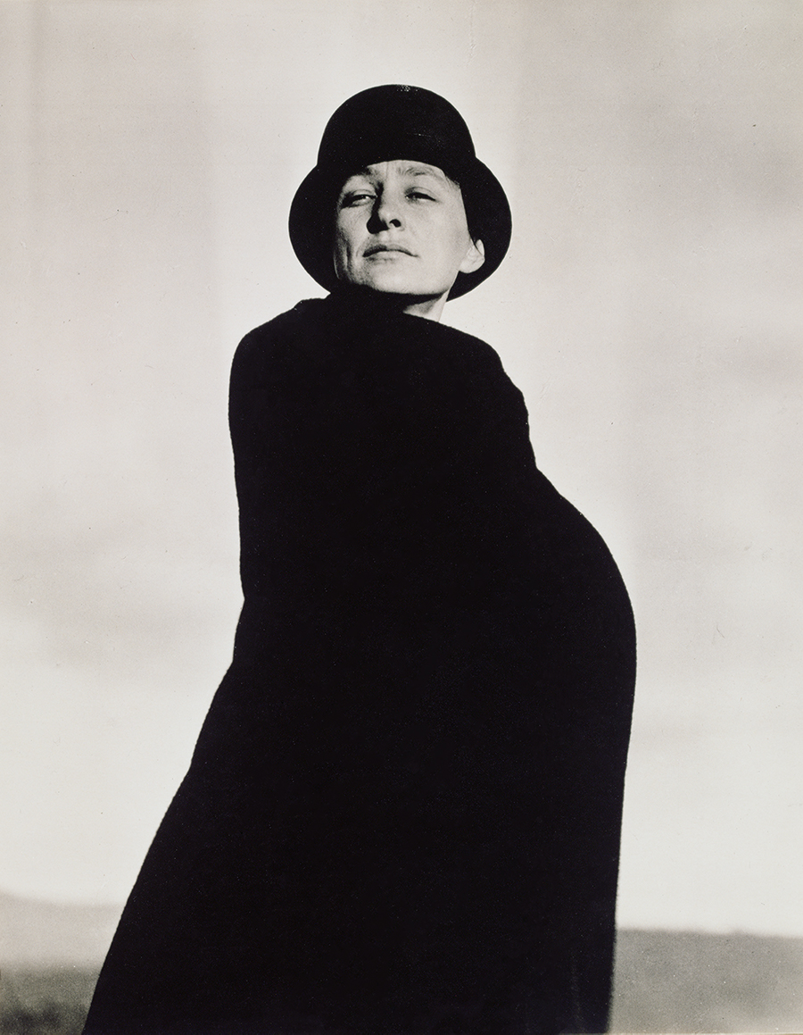 Black and white photograph. We look slightly up at O’Keeffe as she stands swathed in a black coat or wrap, wearing a black hat pulled down to just above her brow, against a white, washed-out sky. Shown from the knees up, her body is angled to our right but she turns her face to us. She gazes down her long nose through narrowed eyes, off to our left. Lit from our right, sharp shadow defines the hollow under her high cheekbone on our left. Her lips are set in a line, and she seems to lean a little away from us. Her entire body is hidden behind her black garment. The light gray horizon comes about an eighth of the way up the composition, and the rest of the background is pale sky.