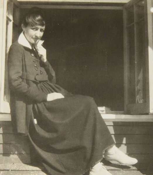 Black and white photograph. O’Keeffe sits on the deep ledge of a window opening and looks at us, one hand raised to the upturned collar of her shirt. The square window opening fills the top three-quarters of the picture, and glass-paned panels open toward us. On our side of the window, O’Keeffe sits on the sill, her crossed legs angled to our right, her back leaning against the left edge of the opening. She turns her oval face to look at us from the corners of her eyes. She pulls her chin back a bit, a faint smile on her lips. Her dark hair is swept loosely up, and it blends with the shadowy room behind her. Her black coat has round buttons down the front and is held loosely in place with a belt. Her long, dark skirt covers her legs to her ankles, above white socks and shoes. Her right hand, to our left and closer to us, rests in her lap. Her other hand is raised to the tall point of her upturned collar, by her left cheek.