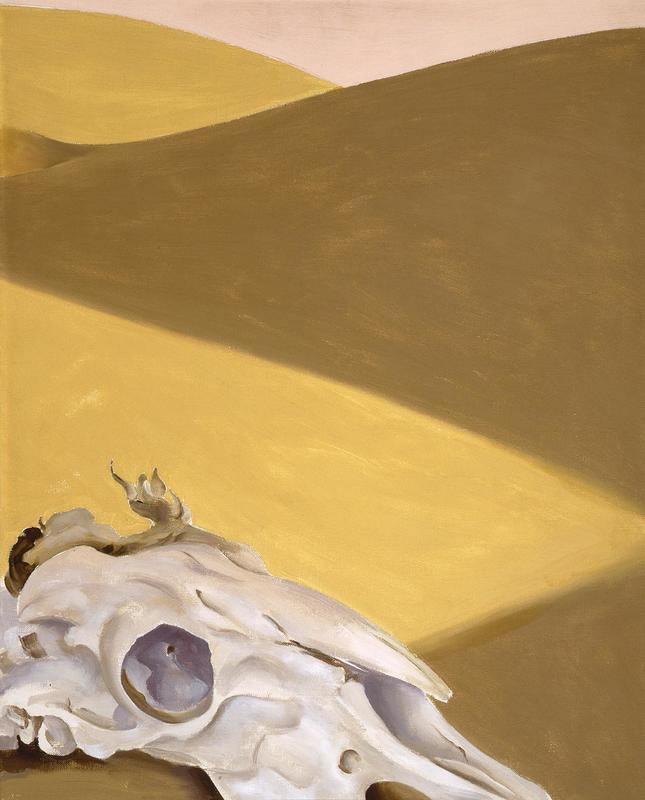 Painting. A white, dry goat’s skull sits on a sandy dune in the lower left corner of this vertical painting. Two dunes rise in the distance beyond, nearly filling the composition. The skull is painted parchment white shaded with pale lavender purple. An undefined, curling tuft near the head could be the remnants of the goat’s skin or fur. The skull is angled to our right, almost in profile. Sun washes the sandy area beyond the skull in golden yellow. The dune that curves up and to our right is darker, army brown. Another sunlit dune fills the top left corner. A sliver of pale pink sky stretches across the top edge of the canvas.